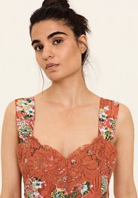Oranje jurk met bloempatronen en kantafwerking. Heeft brede bandjes en een aansluitende corsage. Gestructureerde stof met sequins accenten.
