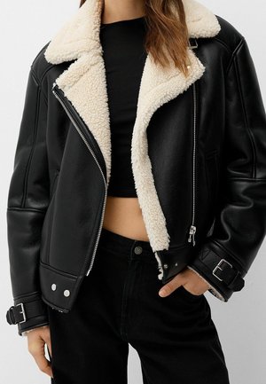 Femme portant une veste de motard en cuir noir avec doublure en peau lainée blanche, un crop top noir et un pantalon noir, main visible dans la poche.