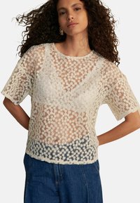 Haut en dentelle blanche avec un motif floral, manches courtes et coupe ample. Présente un design texturé, une superposition semi-transparente et un col rond.