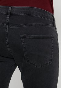 Vaqueros de mezclilla negros con una textura suave, cintura de altura media y un único bolsillo trasero visible. Costuras simples a lo largo de los bordes.