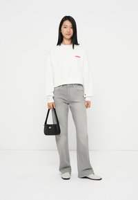 Witte cropped sweatshirt met rood logo, grijze flared jeans, zwarte handtas met logo en beige schoenen. Model staat tegen een witte achtergrond.