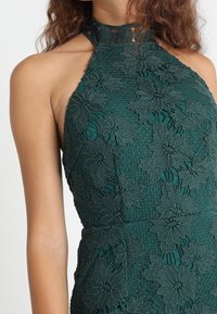 Dunkelgrünes Spitzen-Halterkleid mit floralen Mustern, strukturiertem Stoff und Ausschnitten im Nacken.