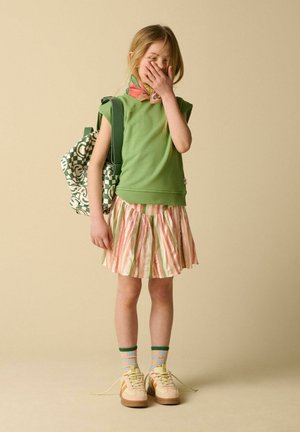 Jeune fille en haut sans manches vert et jupe rayée couvre sa bouche avec la main, portant des chaussettes à motifs, des baskets et un sac à dos.