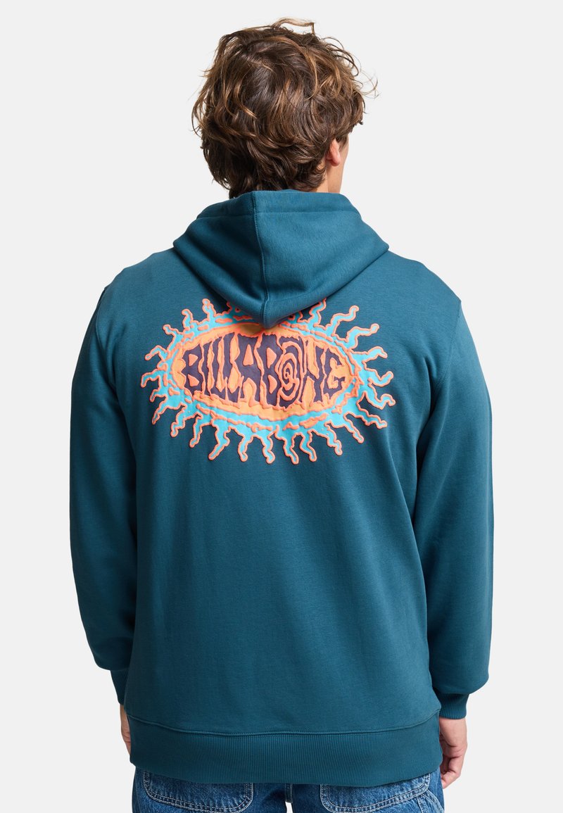 Person trägt einen türkisfarbenen Hoodie mit orange-blauem Sonnenstrahl-Design und dem Schriftzug "Billabong" auf dem Rücken, kombiniert mit blauen Jeans.