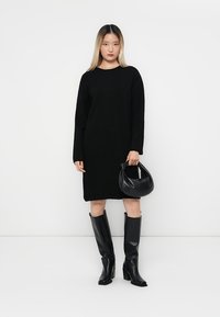 Abito nero lavorato a maglia con maniche lunghe e scollo rotondo. Abbinato a stivali neri al ginocchio e a una piccola borsa nera rotonda.