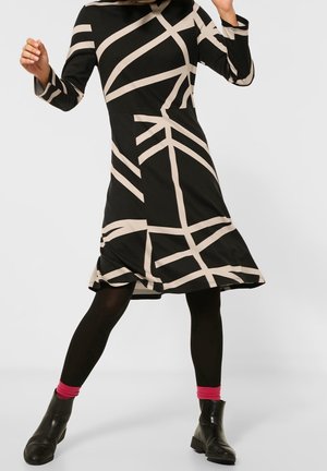 Robe noire avec des lignes géométriques beiges, manches longues, ourlet évasé et fente sur le côté. Portée avec des bottes noires et des chaussettes roses.