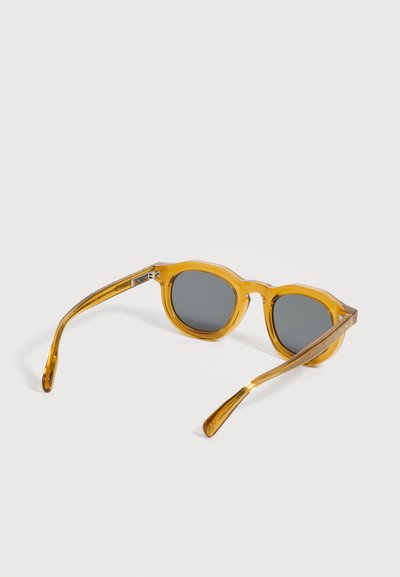 Lunettes de soleil rondes transparentes de couleur ambre avec des verres teintés foncés exposées sur une surface blanche.