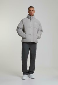 Grå puffjakke med høy krage og quiltete mønster, kombinert med mørke bukser og hvite sneakers. Modellen står med hendene i lommene.