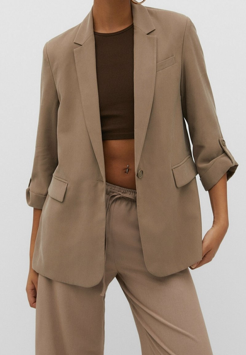 Personne portant un blazer beige avec les manches retroussées, un pantalon assorti avec une taille à cordon, et un top court marron foncé laissant apparaître un piercing au nombril.