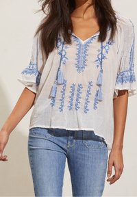 Vit blus med blå broderade mönster, med fransar, V-ringning och korta ärmar; ihop med denimjeans.