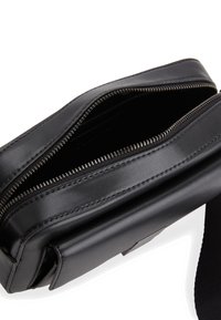 Černá kožená taška se zipovým uzávěrem, obdélníkový tvar, hladký povrch a odnímatelný popruh. Interiér je viditelný a je prázdný.
