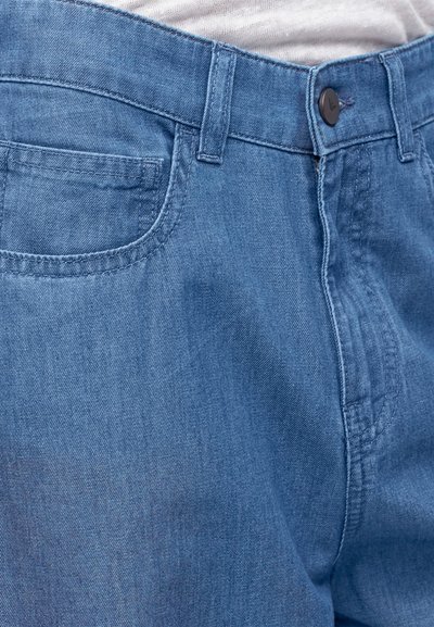 Κοντινή λήψη μπλε τζιν μπροστά με θηλιές για ζώνη, κλείσιμο με κουμπί, μπροστινή τσέπη και λευκό πουκάμισο που έχει μπει στη μέση.