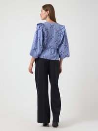 Blouse à carreaux bleus avec imprimé floral, épaules froncées et manches longues, associée à un pantalon large noir et des chaussures à talons.