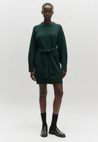 Grünes gestricktes Kleid mit langen Ärmeln, hohem Kragen und einem Stoffgürtel. Das strukturierte Material und der gerippte Saum betonen das Design.
