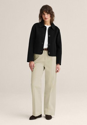 Nainen, jolla on musta napitettu takki, valkoinen paita, beiget korkeavyötäröiset housut, ruskea vyö ja tummanruskeat kengät, seisomassa yksinkertaisen taustan edessä.