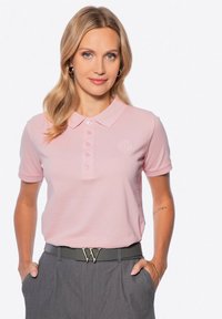 Světle růžové polo tričko s vroubkovanou texturou, límečkem a zapínáním na tři knoflíky. Obsahuje vyšívané logo a je spárováno se šedými kalhotami.