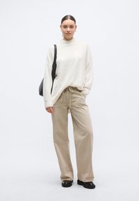 Smėlinės spalvos didelio dydžio megztinis su aukštu kaklu, smėlio spalvos kelnės su raišteliais ir juodi platforminiai batai. Paprastas dizainas, minkšta tekstūra, laisvas prigludimas.