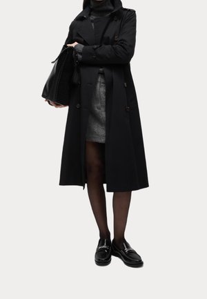 Mujer con abrigo negro, vestido de punto gris, medias negras y mocasines negros, sosteniendo un bolso grande negro.
