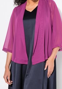 Kvinna iklädd en genomskinlig magenta öppet fram jacka över en svart plisserad klänning, med avslappnade händer vid sidorna mot en vit bakgrund.