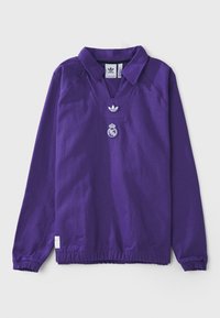 Fialová mikina s límcovým designem, která má výstřih do V a dlouhé rukávy. Má vyšívané logo: Adidas na hrudi a Real Madrid pod ním.