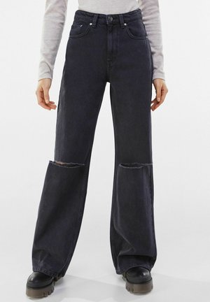 Frau trägt hoch taillierte schwarze Jeans mit weitem Bein und Rissen an den Knien, kombiniert mit schwarzen klobigen Plateau-Stiefeln und einem hellgrauen Langarmshirt.