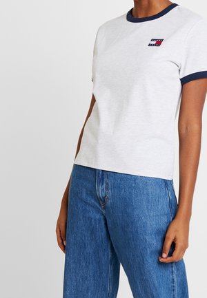 Personne portant un T-shirt blanc avec un col et des bords de manches bleu marine, un petit logo sur la poitrine et un jean bleu taille haute.