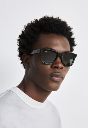 Black Ray-Ban sunglasses with a square frame, dark lenses, and silver hardware accents. Model wears a light gray crew-neck sweater.