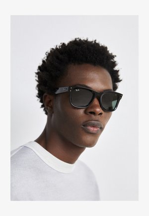 Czarne okulary przeciwsłoneczne Ray-Ban w kwadratowej oprawie, ciemne soczewki i srebrne akcenty. Model nosi jasnoszary sweter z okrągłym dekoltem.