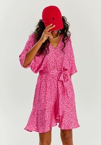Růžové krátké šaty s rozšířeným lemem, zdobené červenými puntíky, s výstřihem do V a stahovacími pásky. Doplněné červenou kšiltovkou a vrstvenými náhrdelníky.