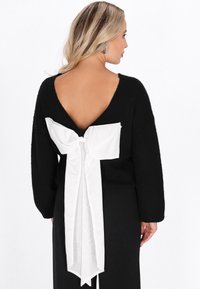 Schwarzer Pullover mit tiefem V-Ausschnitt auf der Rückseite, der ein weißes Stoffschleifendetail in der Mitte aufweist. Das Material wirkt weich und strukturiert.