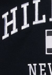 Tessuto nero con testo ricamato bianco, che presenta "HILL" sopra "NEW," accanto a un accento grafico rettangolare in monocromia.