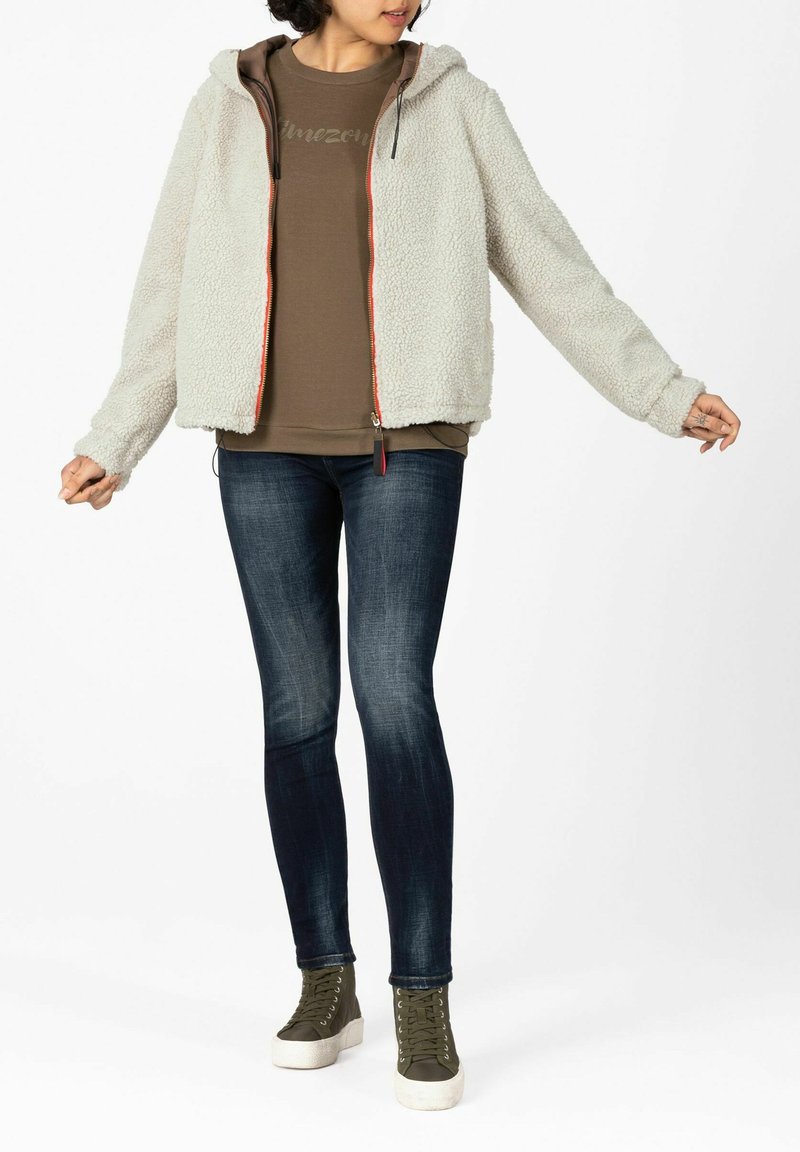 Light gray fleece jacket with hood and orange zipper detail, worn over a brown top, paired with dark blue jeans and green lace-up sneakers.