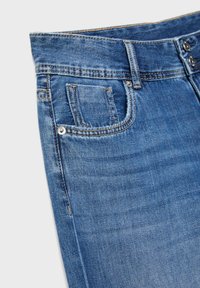 Modré riflové džínsy v zábere zblízka zobrazujú predný vrecko s mincovým vreckom, pútka na opasok, šité švy a detaily z kovových nitov.