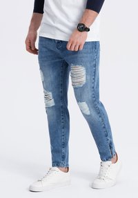 Jeans de mezclilla azul con secciones desgastadas, ajuste entallado y roturas. Combinados con zapatillas blancas y un reloj negro.