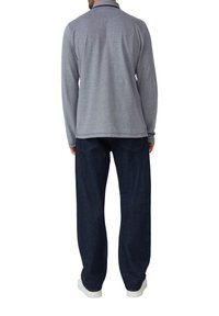 Mann in grauem Langarmshirt, dunklen Jeans und weißen Schuhen, der mit dem Rücken zur Kamera vor einem weißen Hintergrund steht.