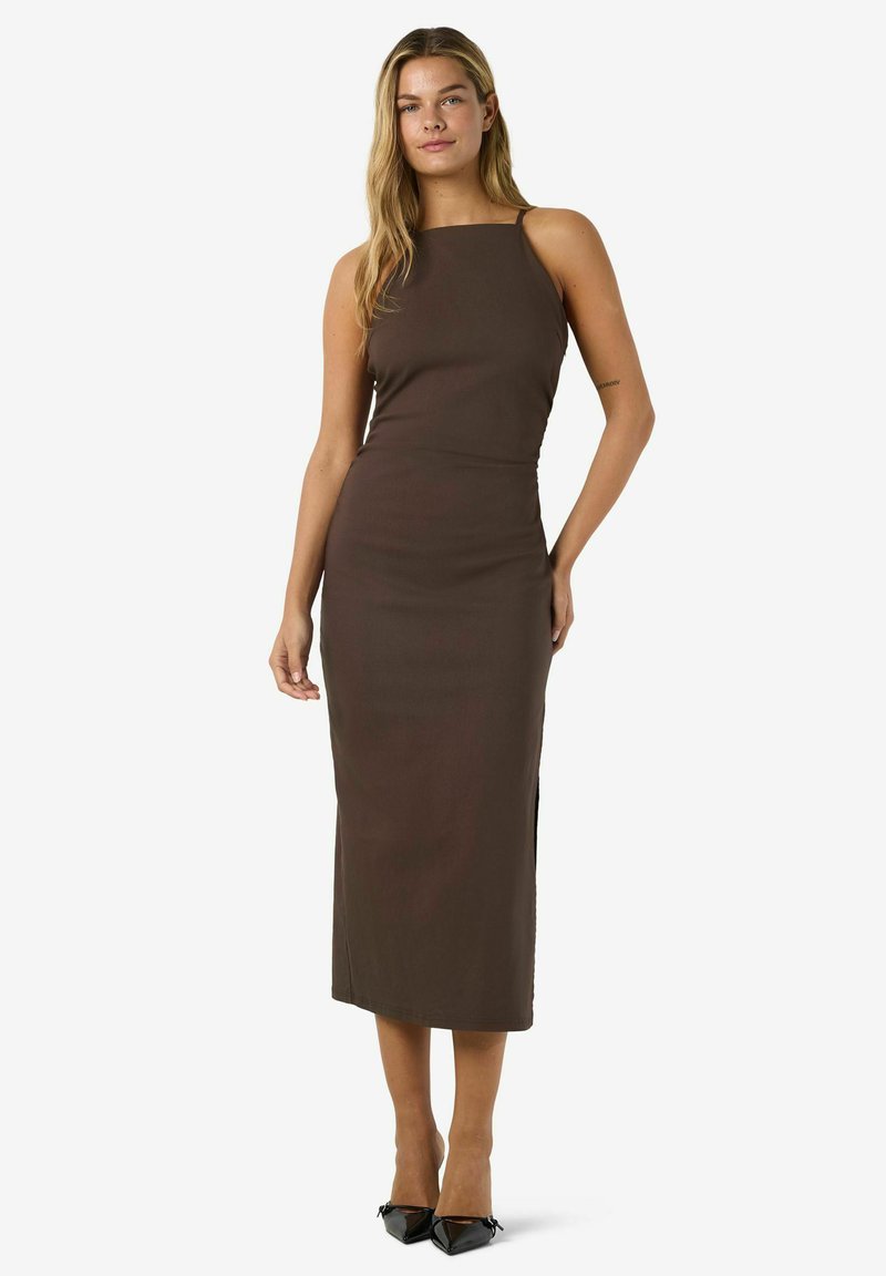Femme debout portant une robe midi sans manches marron foncé avec un décolleté carré et des escarpins noirs pointus sur un fond uni.