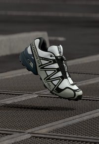 Shoe de corrida leve em branco e preto com parte superior texturizada, marca GORE-TEX e sola aderente para tração.