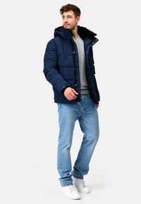 Marineblå pufferjakke med aftagelig hætte, lynlås og quiltet design, parret med en grå sweatshirt og lyseblå jeans.