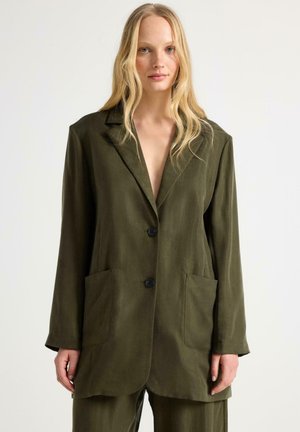 Vaaleahiuksinen nainen, joka on pukeutunut ylisuureen tummanoliivinvihreään bleiseriin, jossa on kaksi nappia ja etutaskut, seisomassa yksivärisen valkoisen taustan edessä.