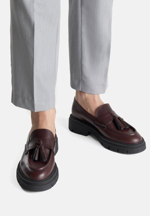 Bordowe mokasyny ze skóry z frędzlami, zaprojektowane z masywną czarną podeszwą. Noszone z jasnoszarymi, dopasowanymi spodniami, podkreślającymi buty.