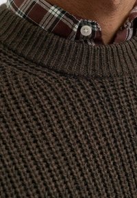 Maglione marrone lavorato a maglia con un design testurizzato e a coste. Sotto indossa una camicia a quadri bordeaux e bianca con colletto e bottoni rotondi.