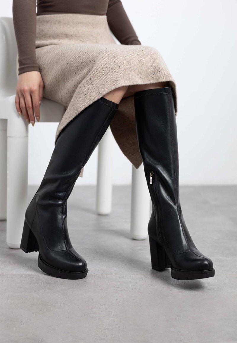 Botas de cuero negro hasta la rodilla con un tacón grueso, cremallera lateral y textura suave, combinadas con una falda de punto beige.