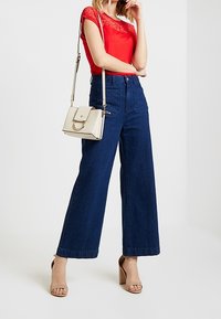 Donna con top in pizzo rosso, jeans a vita alta e gamba larga blu, sandali con tacco beige e borsa a spalla beige con dettagli in oro.