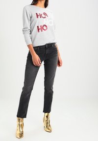 Felpa grigia con scritta "HO HO HO" in paillettes rosse, abbinata a jeans neri e stivaletti alla caviglia color oro metallizzato. Modello in piedi su uno sfondo semplice.