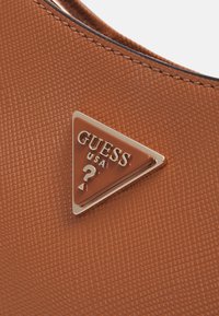 Brązowy materiał o fakturze z trójkątnym metalowym logo z napisem "GUESS USA" i znakiem zapytania, przymocowany do torebki lub akcesorium.