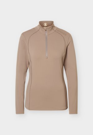 Mujer de pie mirando hacia adelante con una camiseta beige de manga larga con cremallera, pantalones a juego con cinturón y zapatillas blancas sobre un fondo liso.