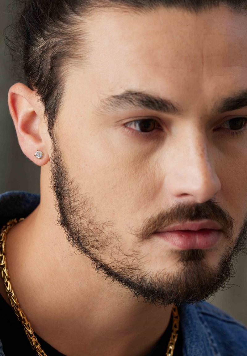 Zilveren studoorbel aan het linkeroor, close-up van een mannengezicht, met kort donker haar, een baard en een gouden ketting om de nek.