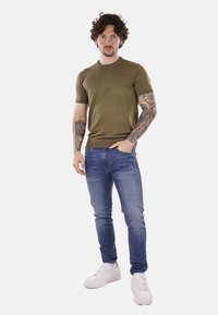 Maglietta verde oliva a maniche corte, jeans aderenti blu e sneakers bianche. Caratteristiche includono tatuaggi sul braccio e vestibilità rilassata.