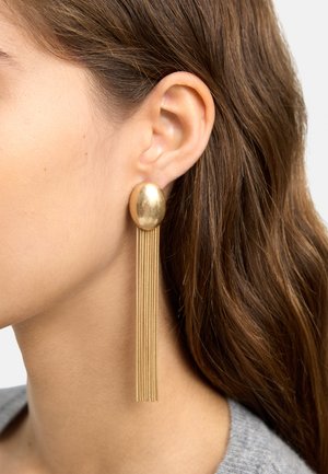 Zbliżenie na ucho kobiety noszącej duży owalny złoty kolczyk z wieloma cienkimi złotymi łańcuszkami zwisającymi w dół.