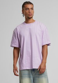 T-shirt oversized lilas avec des rayures blanches verticales et un petit logo brodé sur la poitrine. Tissu en coton doux, coupe décontractée.